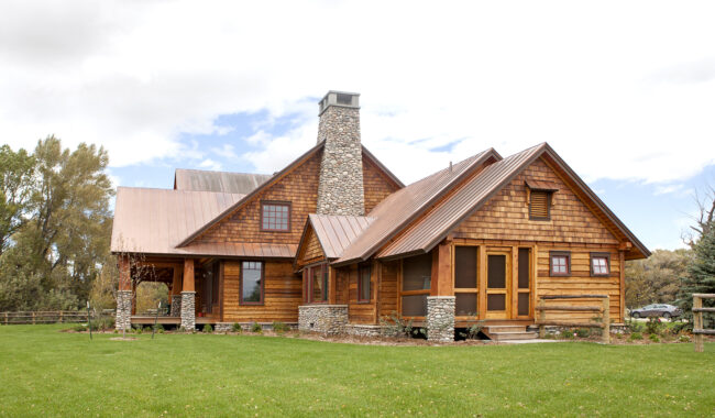 A rustic wooden house with a stone chimney sits on a green lawn under a partly cloudy sky, surrounded by trees and a wooden fence.