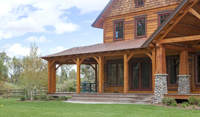 A rustic wooden house with a wraparound porch, stone pillars, and a green lawn in front. Outdoor furniture is on the porch, and trees and a wooden fence are visible in the background under a partly cloudy sky.