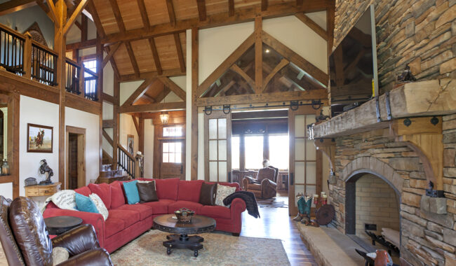 Spacious rustic living room with exposed wooden beams, stone fireplace, red sectional sofa, wooden furniture, and large windows letting in natural light. A loft area and staircase are visible in the background.