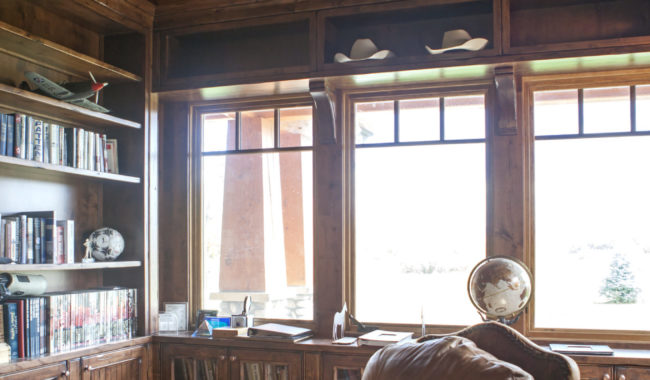 A cozy study with wooden shelves, books, and globe near a large window. An armchair with a striped blanket draped over it sits on a wooden floor, under exposed wooden beams and a slanted ceiling.