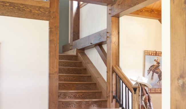 Wooden staircase with intricate carvings leads to an upper level in a rustic home with exposed beams. A cowboy-themed poster and hats hang on the wall, and woven baskets sit by the stairs on a light wood floor.