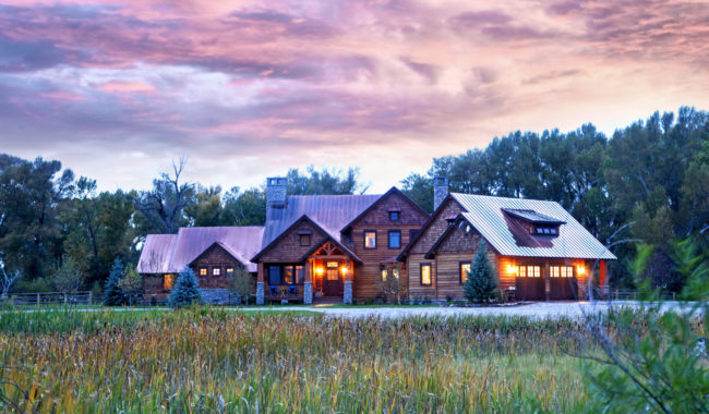 A rustic wooden house with glowing windows sits amid tall grass and trees under a vibrant, colorful sunset sky, creating a peaceful rural scene.