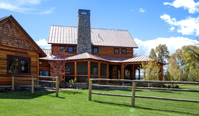 A modern rustic wooden house with a stone chimney, metal roof, and large porch sits behind a wooden fence on a green lawn under a blue sky with scattered clouds. Trees are visible in the background.