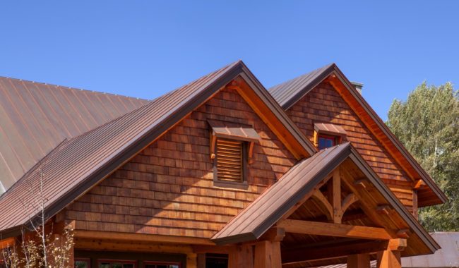 A close-up of a wooden house with a shingle roof and brown siding, featuring triangular gables and a small louvered vent, set against a clear blue sky and surrounded by trees.