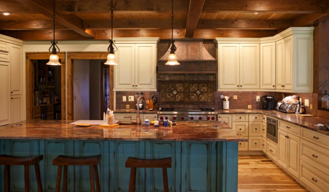 Rustic kitchen with cream cabinets, a large wooden island with teal base and stools, pendant lights, wooden ceiling beams, and a range with a decorative hood. Warm lighting and wooden floors create a cozy atmosphere.