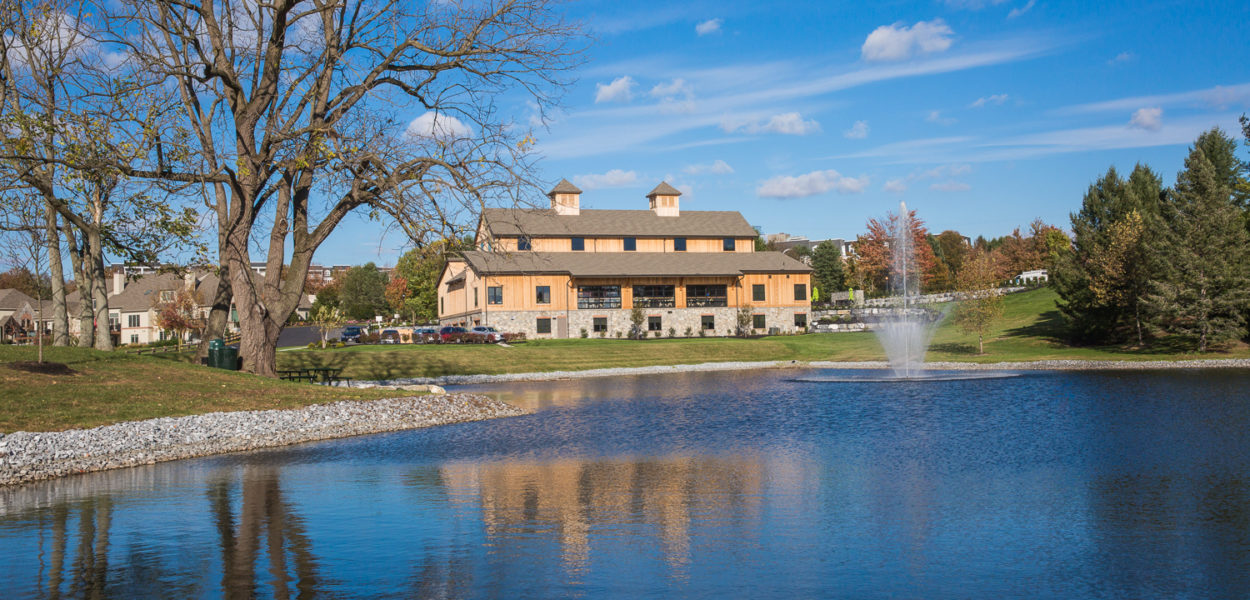 A large building with tan walls and stone accents sits behind a pond with a fountain, surrounded by grass, trees, and blue sky with scattered clouds. The building and trees are reflected in the water.