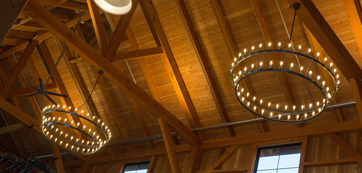 Wooden ceiling with exposed beams, two large circular chandeliers with many small lights, an industrial pendant light, and windows letting in natural light.