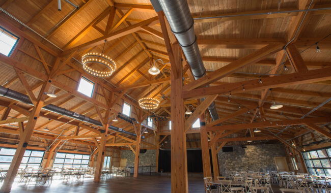 A spacious hall with exposed wooden beams, circular chandeliers, large windows, stone walls, and scattered tables and chairs, creating a rustic and airy atmosphere.