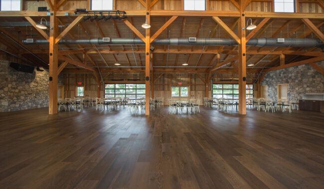 Spacious rustic event hall with wooden beams, high ceiling, exposed ductwork, stone walls, large windows, and empty dark wood floor. Round tables and chairs are arranged along the walls.