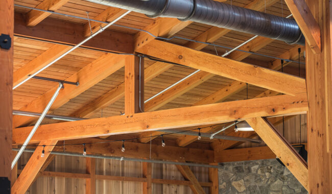 Exposed wooden beams and metal ductwork on the ceiling of a building, with part of a stone wall visible below. The structure and joinery of the wood are highlighted in natural light.
