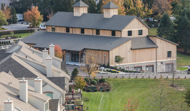 A large wooden barn-style building with two cupolas, surrounded by green lawns and trees with autumn foliage, sits near a row of townhouses in a suburban neighborhood.