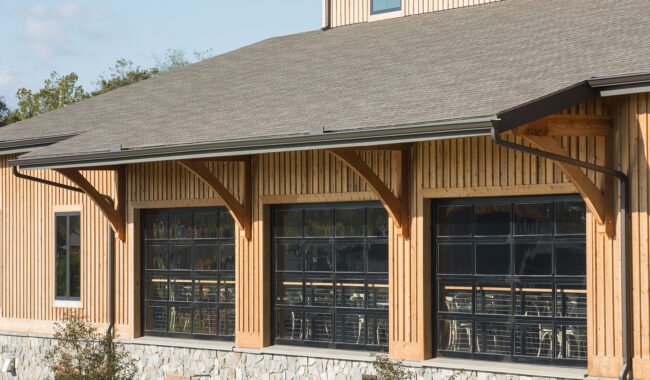 A wooden building with large glass garage-style doors and a stone foundation, featuring exposed wooden beams under the roof overhang. Tables and chairs are visible inside through the windows.