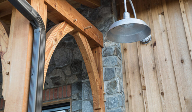 A close-up of a rustic building exterior shows a wooden beam, stone wall, vertical wood siding, and a metal wall-mounted outdoor light fixture.