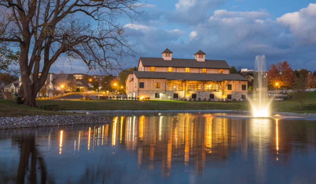 A large building with warm lights sits beside a pond with a bright fountain, reflecting in the water at dusk. Leafless trees and a cloudy sky frame the peaceful scene.