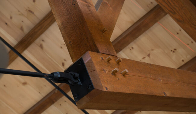 Close-up of a wooden beam joint with metal brackets and wooden pegs, attached to a light fixture arm, set against a wooden ceiling with exposed beams.