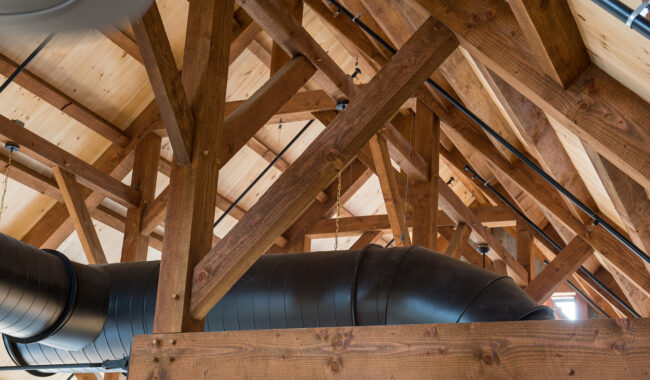 Exposed wooden beams and trusses support a vaulted ceiling with light wood paneling. Large black ventilation ducts run through the open structure, creating a modern industrial look.