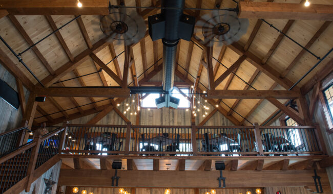 Interior of a rustic building with a high wooden ceiling, exposed beams, industrial fans, hanging lights, and a loft area with tables and chairs above the main floor. Warm lighting creates a cozy atmosphere.