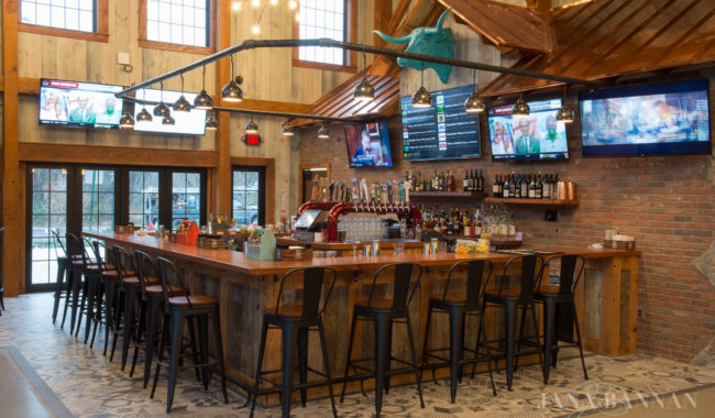 A rustic bar with wooden stools, a large central counter, multiple TVs on the walls, shelves of liquor bottles, and large windows letting in natural light. A turquoise bull skull decorates the wall above the bar.