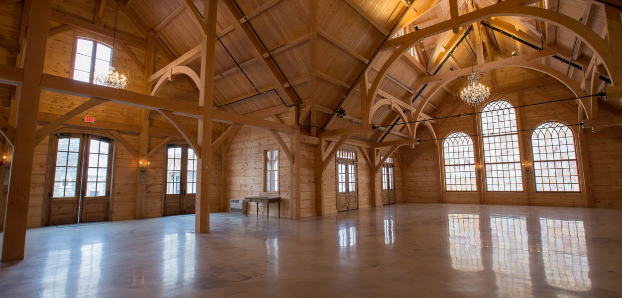 Spacious wooden hall with high vaulted ceilings, large arched windows, exposed beams, chandeliers, and polished floor. Sunlight streams through the windows, illuminating the empty interior.
