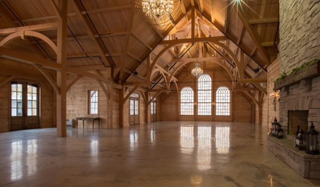 Spacious wooden hall with exposed beams, arched windows, chandeliers, polished floor, and a stone fireplace with decorative lanterns. Natural light fills the rustic, elegant interior.