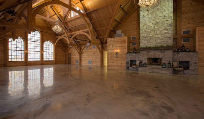 Spacious wooden hall with arched beams, large windows, stone fireplace, chandeliers, and polished floor. The room is empty and has a rustic, elegant ambiance.