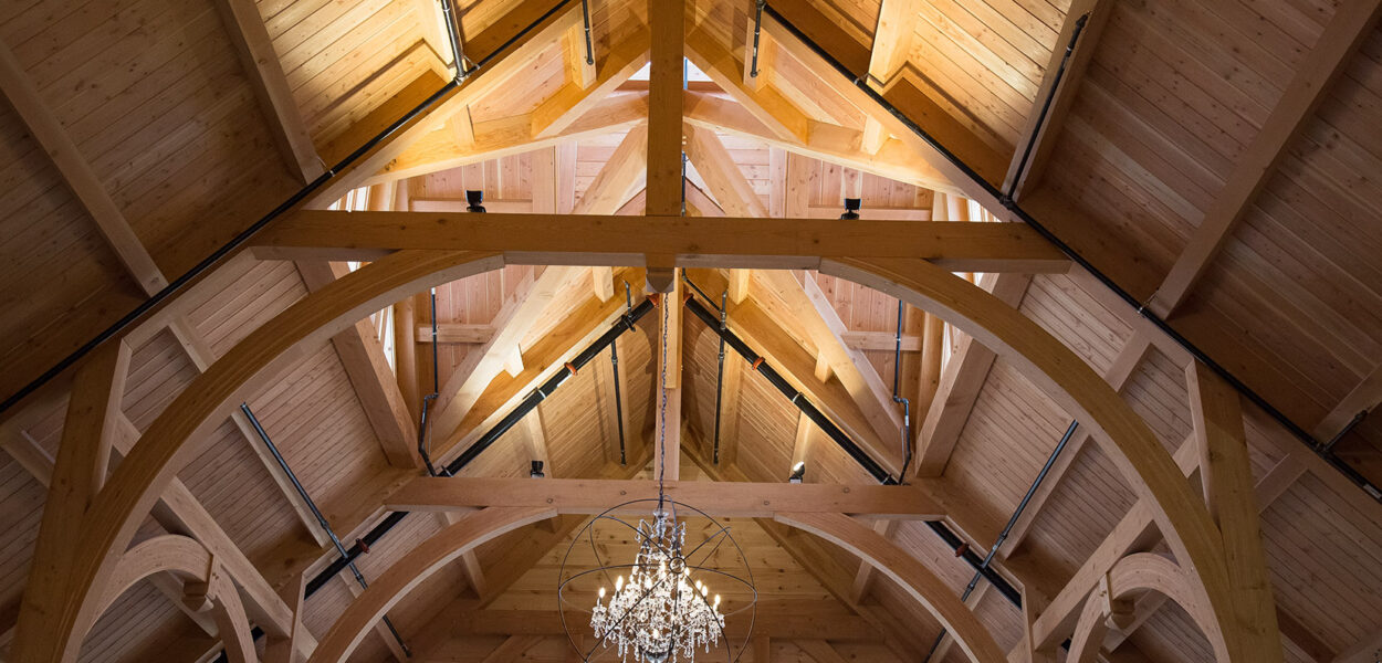 Wooden vaulted ceiling with exposed beams, a central triangular truss, and a decorative chandelier hanging in the middle. Natural light shines through an arched window below.