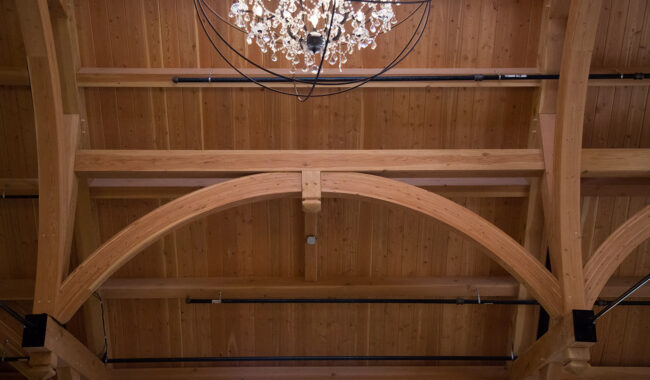 A close-up view of a wooden ceiling with exposed beams and a crystal chandelier hanging down, partially visible at the top of the image. Black pipes run along the beams.