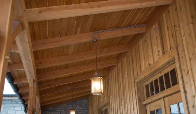 A rustic wooden porch ceiling with exposed beams, two hanging lantern lights, and wood-paneled walls beside a stone wall and wooden-framed windows.