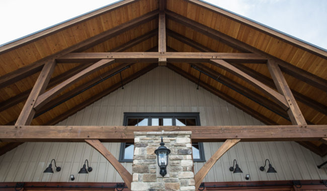 The image shows the gabled roof of a rustic building with exposed wooden beams, a stone column, a black lantern, and beige vertical siding under a blue sky.
