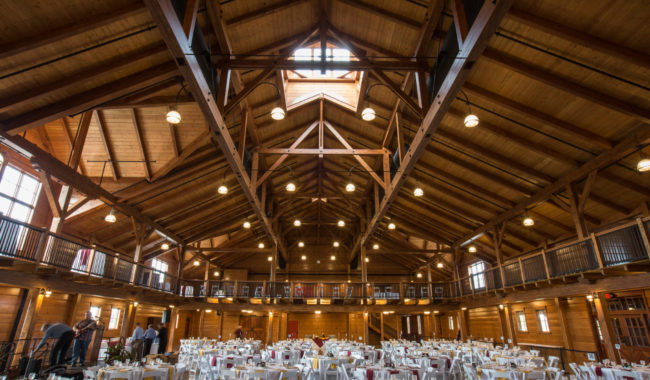 A spacious wooden hall with a high vaulted ceiling, exposed beams, and large windows. Numerous round tables with white chairs are set up for an event, while a few people are standing on the left side of the room.