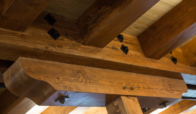 Close-up view of an exposed wooden beam ceiling with large, polished timber beams and metal brackets, showcasing detailed wood grain and craftsmanship in a rustic architectural style.