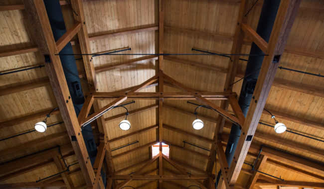 A view looking up at a wooden ceiling with exposed beams, metal supports, and hanging round lights. Skylight windows let in natural light. The structure has a warm, rustic appearance.