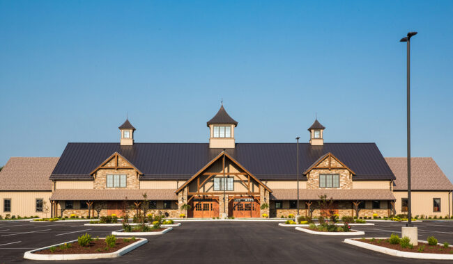 Large, barn-style building with a dark metal roof and three cupolas, stone and timber accents, symmetrical design, and landscaped entrance, situated in an empty parking lot under a clear blue sky.