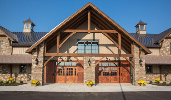 A large, rustic barn with stone and wood exterior, tall peaked roof, wooden double doors, and small cupolas. Yellow flowers in planters line the entrance under a clear blue sky.
