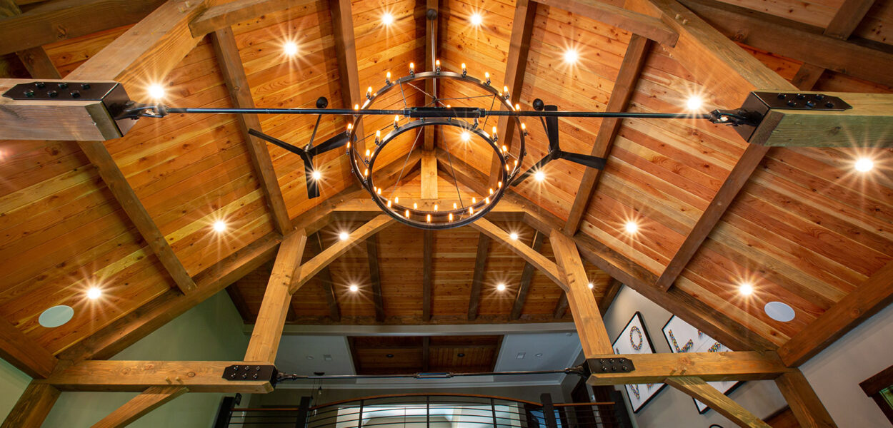 View of a vaulted wooden ceiling with exposed beams, a circular metal chandelier with many lights, and a loft railing beneath, giving a warm and rustic feel to the interior space.