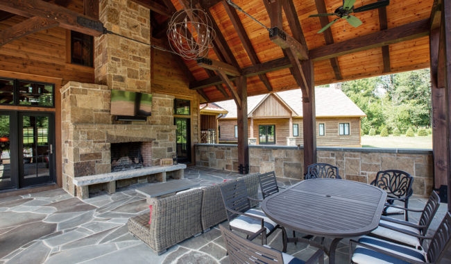 A covered outdoor patio features a stone fireplace with a TV above it, wicker seating, a dining table with chairs, ceiling fans, and wood beams under a gabled roof. Large windows and stone walls open to a view of a house and greenery.