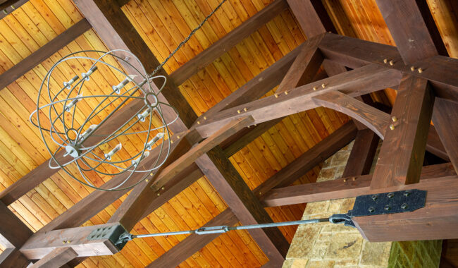 A view looking up at a wooden vaulted ceiling with exposed beams, a metal chandelier with an orbital design, and part of a stone wall visible below.