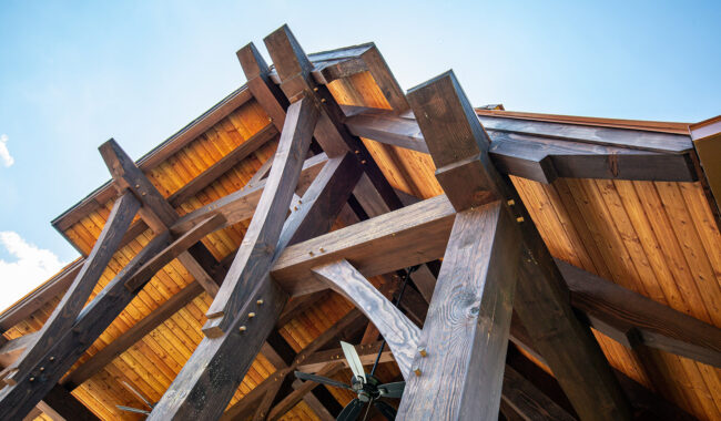 View from below of a timber-framed roof structure with exposed wooden beams and a ceiling fan, set against a clear blue sky. The natural wood and sturdy beams highlight rustic architectural details.