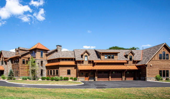 Large, rustic-style log mansion with multiple stories, stone accents, and several dormer windows, surrounded by landscaped greenery and a spacious driveway, under a clear blue sky.