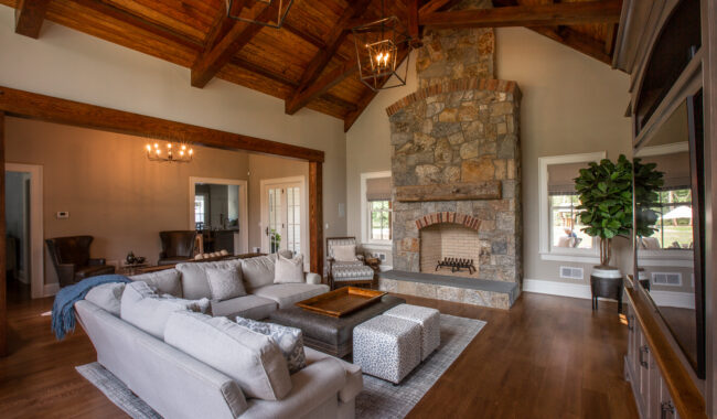 Spacious living room with high wood-beamed ceiling, large stone fireplace, gray sectional sofa, armchairs, coffee table, ottomans, hardwood floors, modern lighting, and a potted plant near a sunlit window.