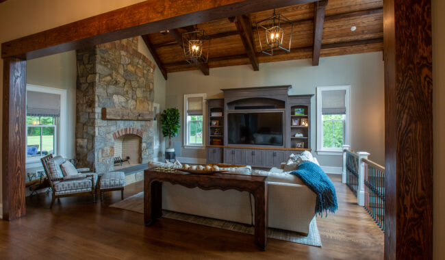 A cozy living room with a wooden-beamed ceiling, stone fireplace, large gray entertainment center, two armchairs, a sofa with blue accents, and hardwood floors. Natural light comes through windows on both sides.
