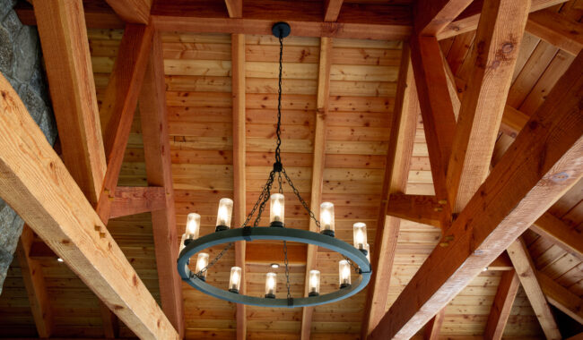 A circular chandelier with candle-style lights hangs from a high ceiling made of exposed wooden beams and planks. The warm wood and soft lighting create a rustic, cozy atmosphere.