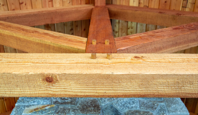 Close-up of wooden beams joined with a traditional Japanese woodworking technique, showing interlocking joints; stone flooring and wood-paneled walls are visible in the background.