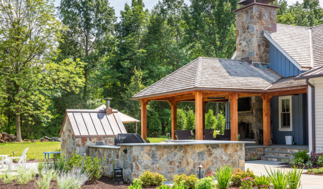 A stone outdoor kitchen and patio with a wood roof, a built-in grill, pizza oven, fireplace, bar seating, and lush landscaping surrounded by green trees on a sunny day.