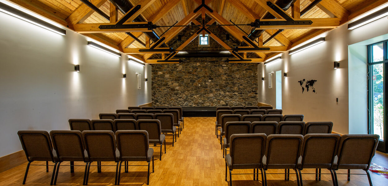 A small chapel with rows of brown cushioned chairs facing a stone wall and stage, wooden vaulted ceiling with exposed beams, and large windows letting in natural light.