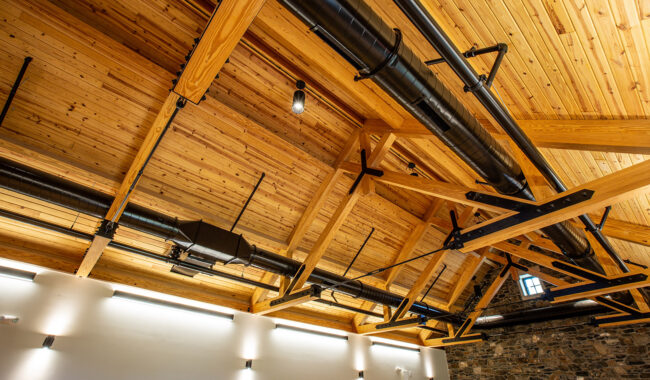 Exposed wood beam ceiling with black metal ducts and light fixtures in a modern building. The ceiling contrasts with stone walls and white upper walls, creating an industrial yet warm atmosphere.