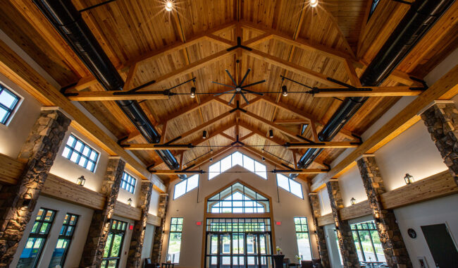 Spacious lobby with high vaulted wooden ceiling, exposed beams, large black ceiling fan, stone pillars, windows on all sides, and double glass doors letting in natural light.