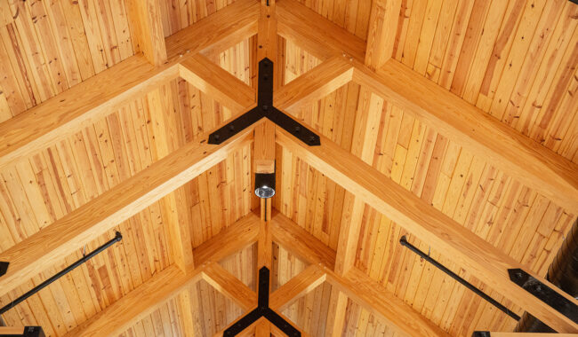Wooden vaulted ceiling with exposed beams and black metal supports, featuring a central light fixture. Warm-toned wood panels create a geometric pattern.
