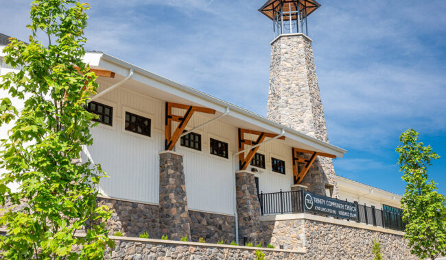 A stone and wood building with a tall bell tower stands under a blue sky. The sign reads Trinity Community Church with service times listed. Green trees frame the foreground.