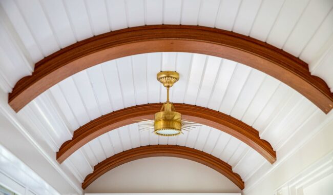 A ceiling with white curved wooden panels and three arched wooden beams. A brass light fixture with a circular, spiked design hangs from the center beam. Natural light enters from windows on the left.